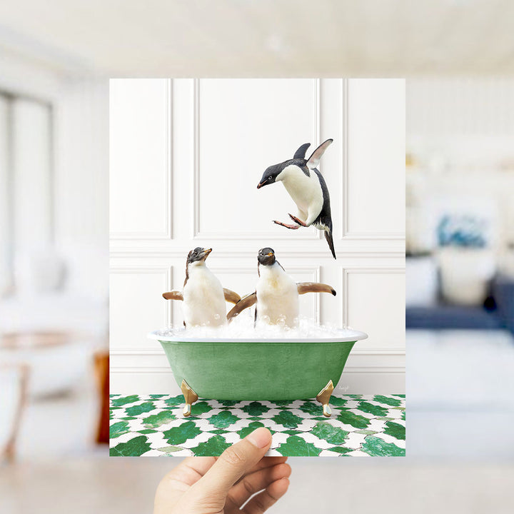 A hand holds up a framed image of two penguins sitting in a green bathtub, with one penguin flying towards the other.
