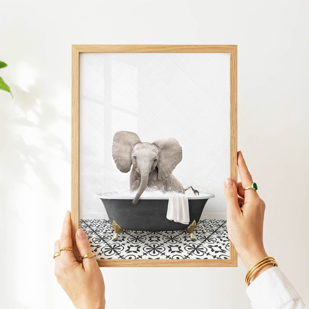A person's hands are holding a framed picture of an elephant sitting in a bathtub filled with water.