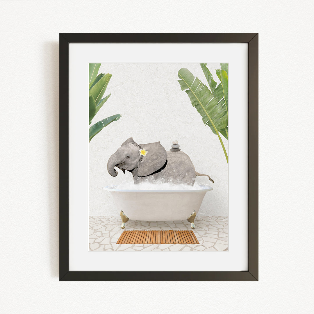 A framed image depicting a gray elephant sitting in a white bathtub, surrounded by green leaves and a wooden mat on the floor.