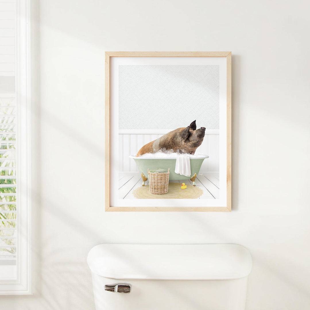A framed image of a pig sitting in a bathtub, surrounded by a basket of bath toys and a towel, is displayed on a white wall above a white toilet.