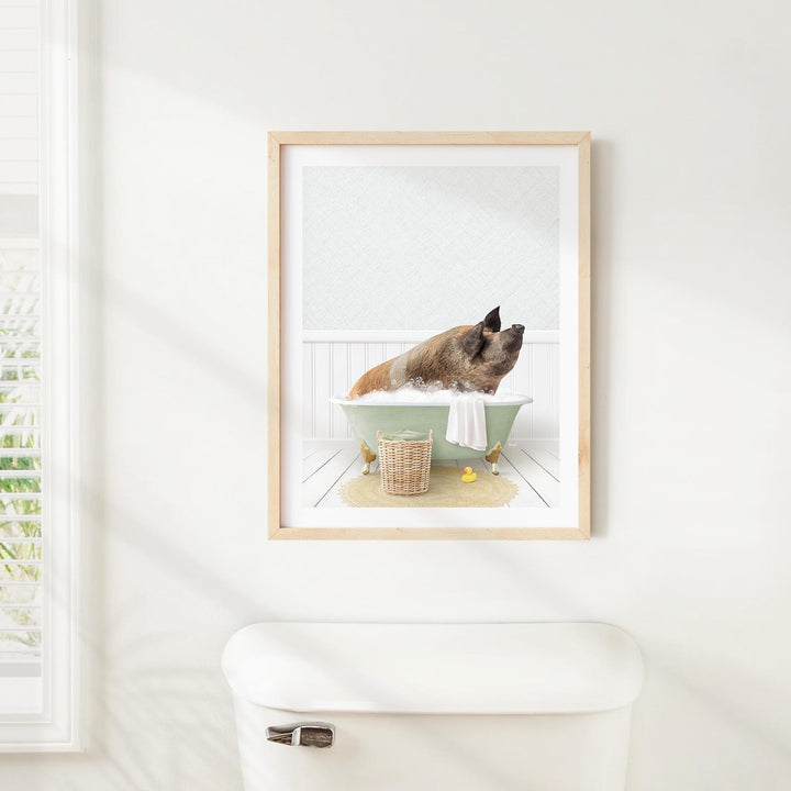 A framed image of a pig sitting in a bathtub, surrounded by a basket of bath toys and a towel, is displayed on a white wall above a white toilet.