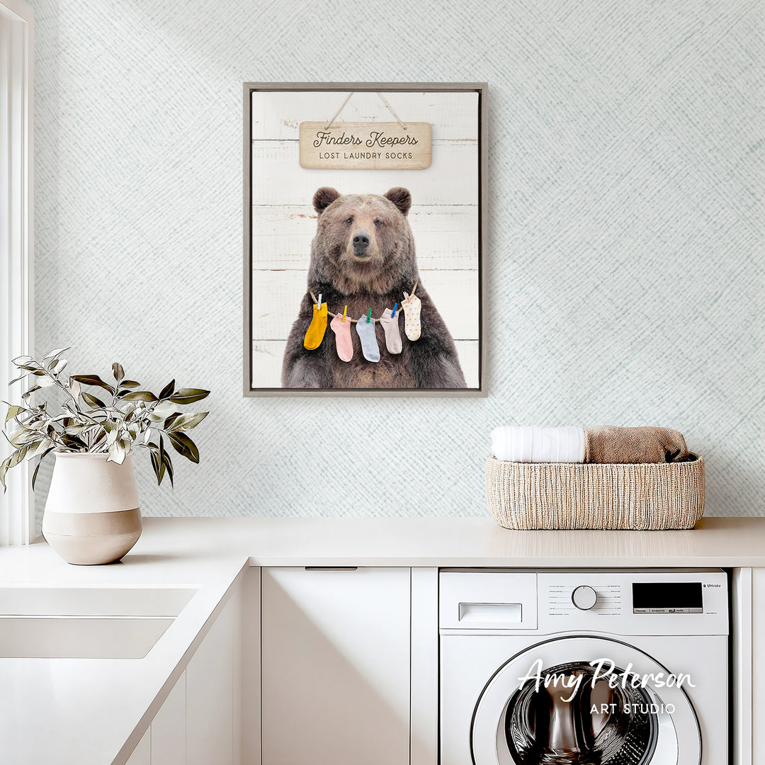 a framed print of a bear hanging laundry on a wall, with a basket of towels and a plant nearby.