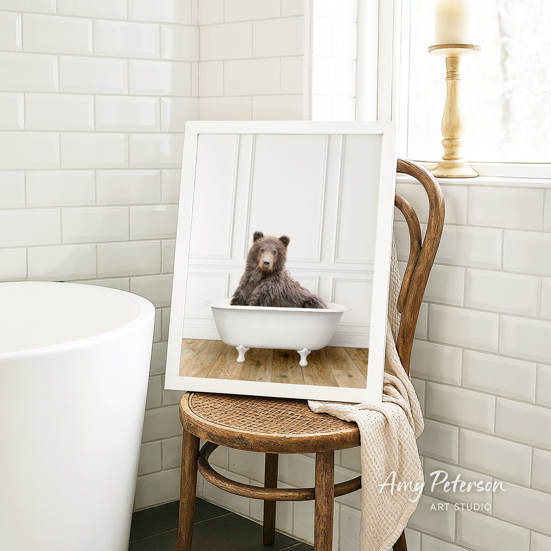 A framed image of a bear sitting in a bathtub is displayed on a wooden chair in a bathroom setting.
