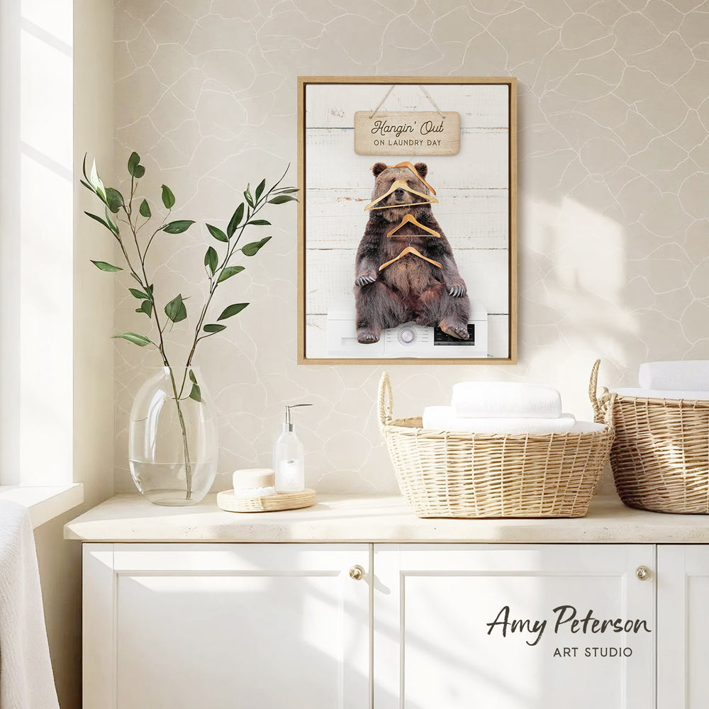 a framed picture of a bear wearing a sign that says "hangin' out on laundry day" hanging on a wall above a white cabinet.