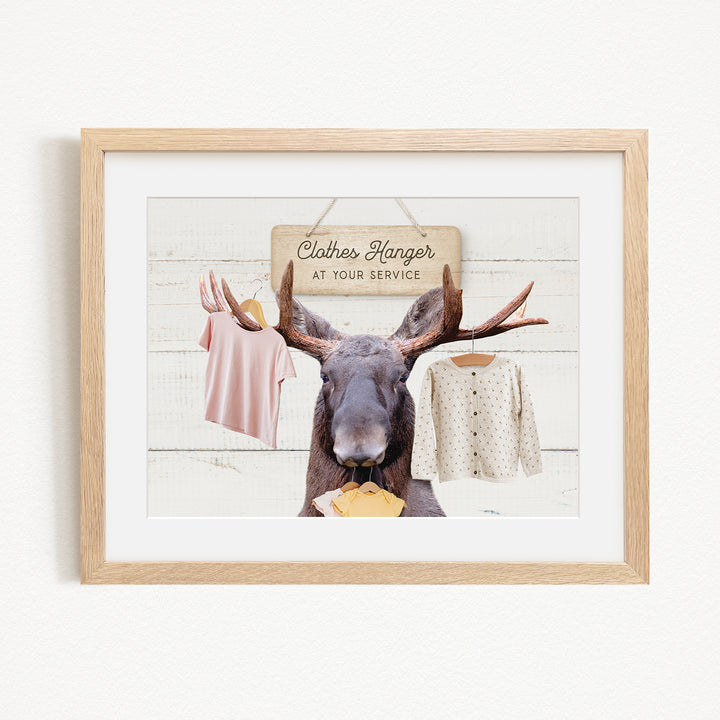 A framed image depicting a moose with antlers holding clothes on a clothesline, with a sign that reads "Clothes Hanger at your Service".