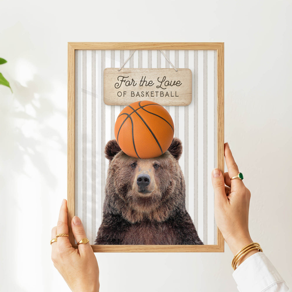 A person's hands are holding a framed picture of a bear with a basketball on its head, which is displayed on a wall.