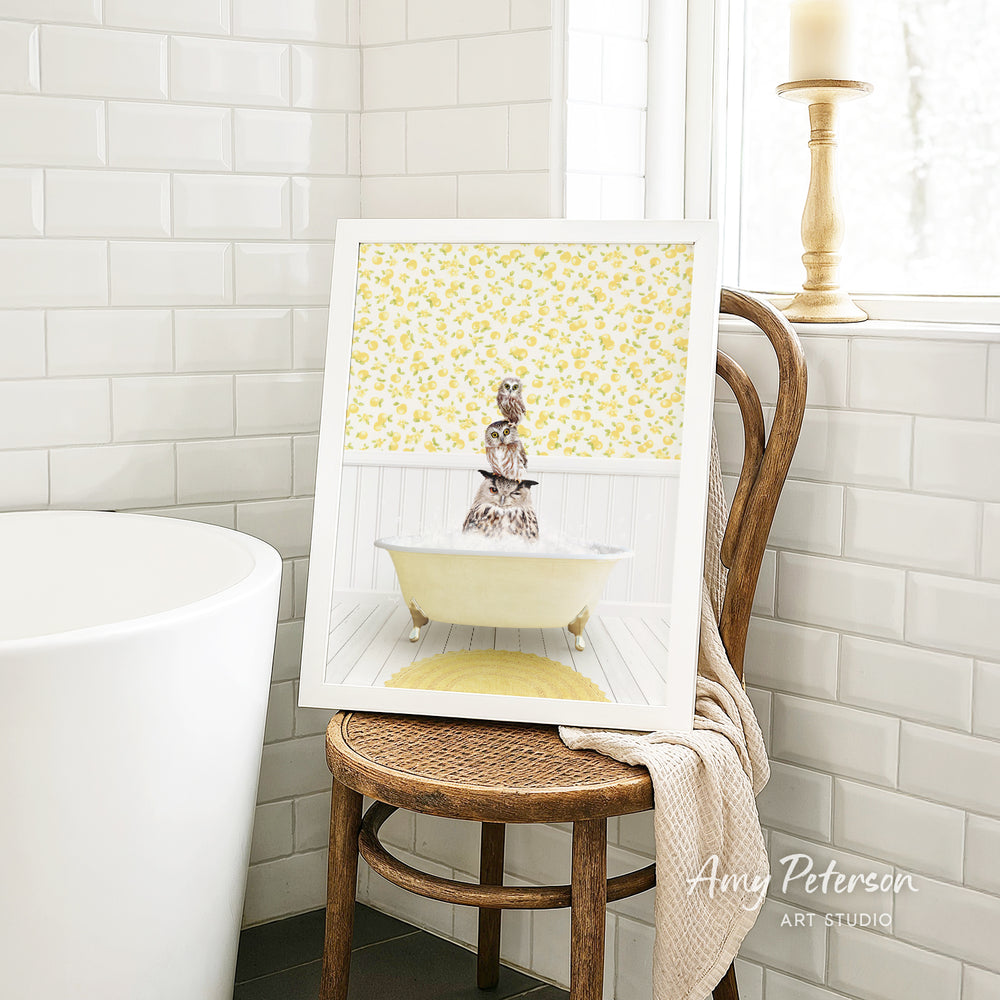A framed image of a cat sitting in a bathtub, surrounded by yellow flowers, is displayed on a wooden chair in a bathroom setting.