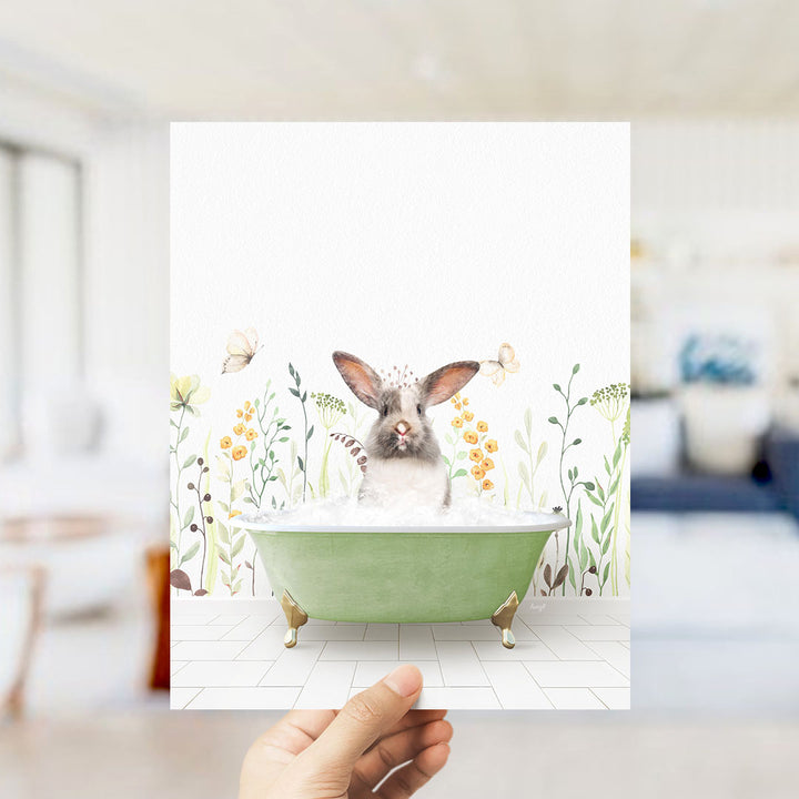 A hand holds up a picture of a rabbit sitting in a green bathtub filled with bubbles, surrounded by a floral background.