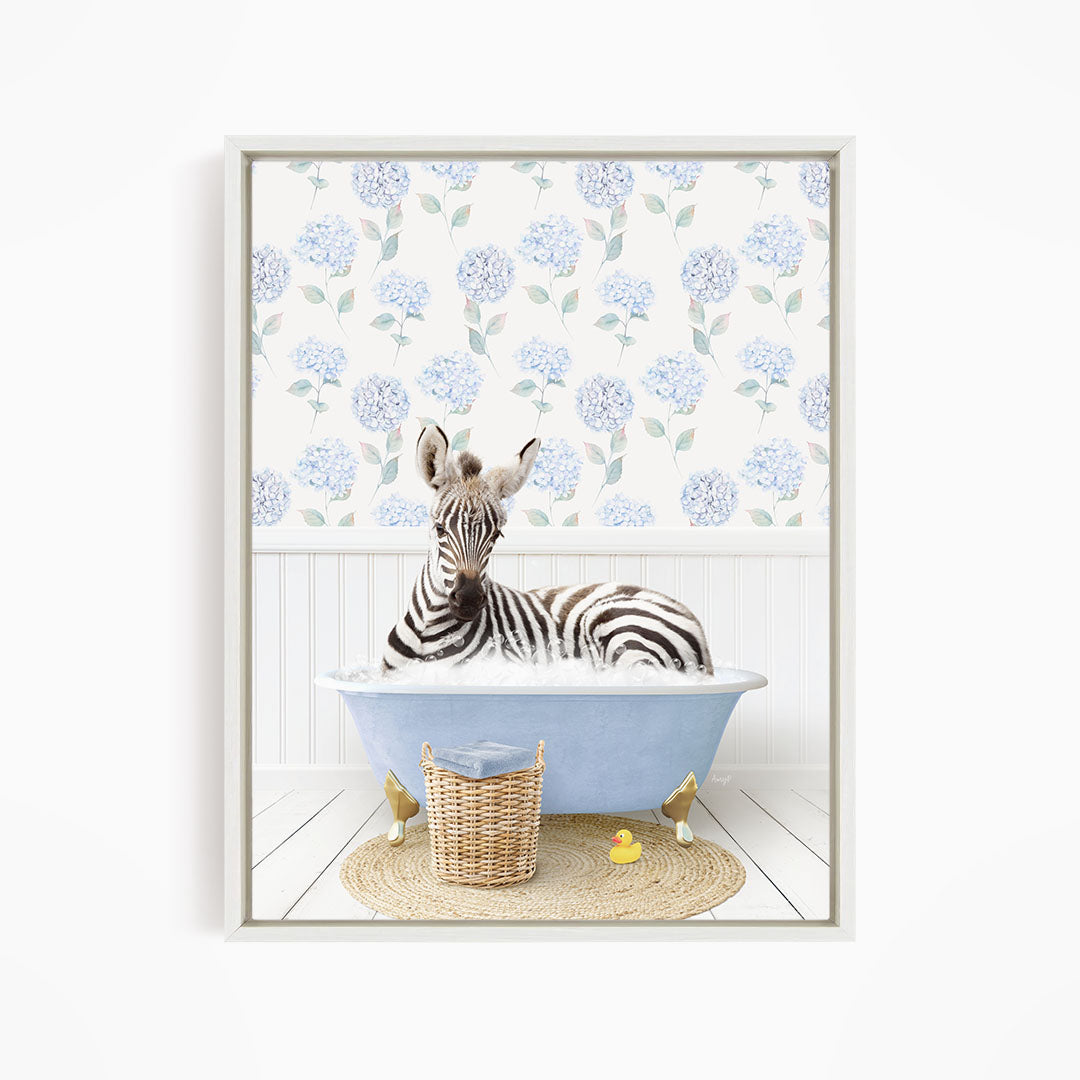 A zebra is sitting in a blue bathtub, surrounded by a wicker basket and a yellow rubber duck, with a floral patterned wall behind it.