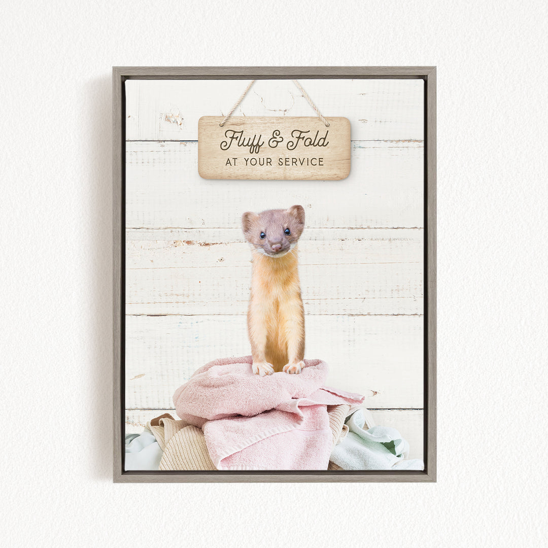 A small brown and white ferret sitting on a pile of clothes, with a wooden sign above it that says "Fuzz & Fold at your Service".