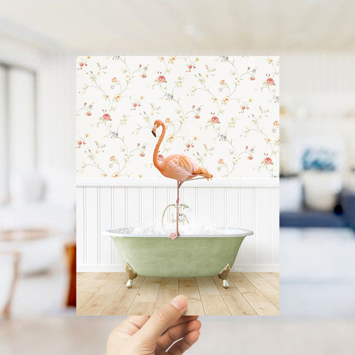 A hand holds up a framed image of a pink flamingo standing on a green bathtub filled with water, set against a floral wallpaper backdrop.