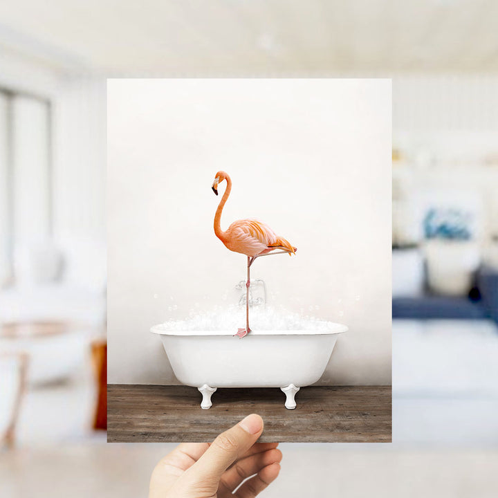 A hand holds up a framed image of a flamingo standing on a white bathtub filled with water, creating a whimsical and playful scene.