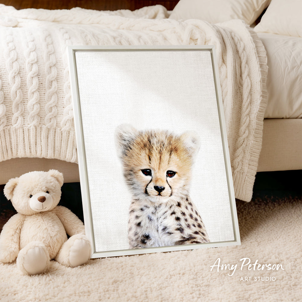 A framed print of a cheetah's face is displayed on a white surface, with a teddy bear sitting next to it.