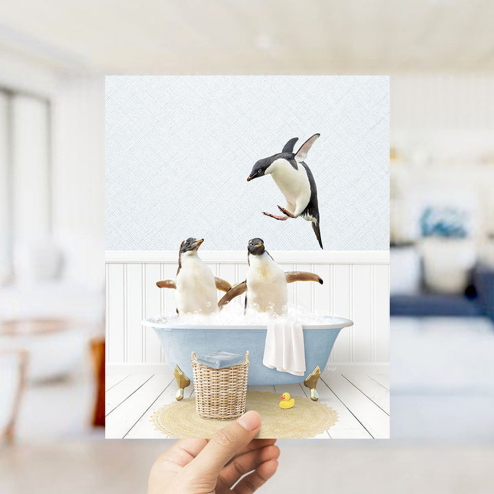A hand holds up a framed image depicting two penguins sitting in a bathtub, with one penguin flying towards the other.